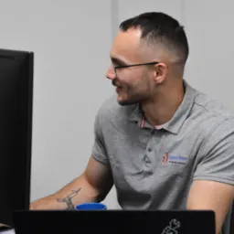 Mężczyzna w okularach, z tatuażem origami na przedramieniu, uśmiecha się patrząc na monitor komputera w biurze, ubrany w szarą koszulkę polo z logo firmy IT.