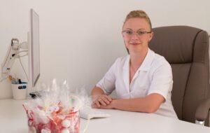 Uśmiechnięta kobieta w okularach i białym uniformie siedzi za biurkiem w jasnym gabinecie. Na biurku widoczny komputer i dekoracyjny kosz z kosmetykami lub produktami do pielęgnacji.