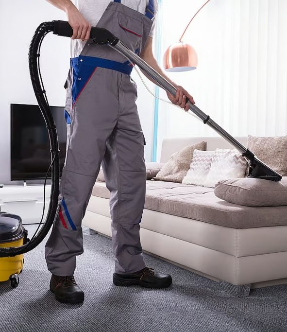 Pracownik w szarym kombinezonie z niebieskimi wstawkami czyści beżową kanapę przy użyciu odkurzacza piorącego z żółtym zbiornikiem, widoczny również fragment szarej wykładziny i telewizora.