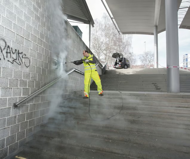 Pracownik w jaskrawożółtym kombinezonie usuwa graffiti z ceglanej ściany przy schodach za pomocą myjki ciśnieniowej, w tle widoczny pojazd serwisowy z otwartym bagażnikiem.