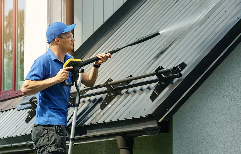 Pracownik w niebieskiej koszulce i czapce, w okularach ochronnych, myje metalowy dach myjką ciśnieniową, stojąc na aluminiowej drabinie z zabezpieczeniem na rynnie.
