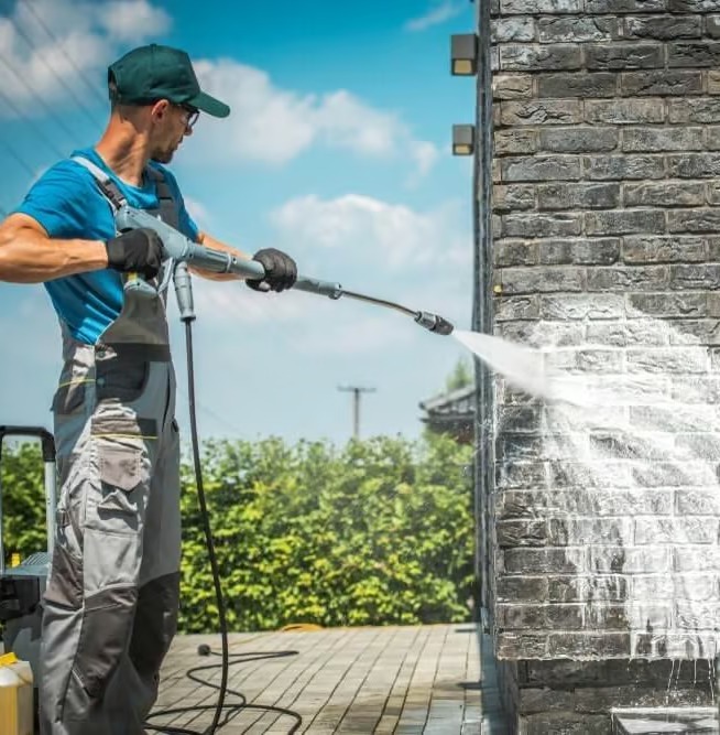 Pracownik w okularach i zielonej czapce czyści elewację z ciemnej cegły za pomocą myjki ciśnieniowej, widoczny efekt czyszczenia.