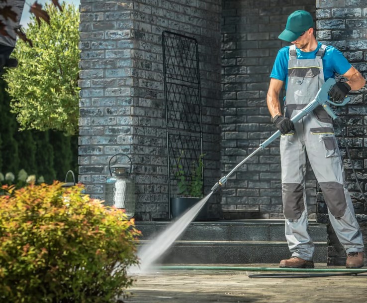 Pracownik w szarym kombinezonie i zielonej czapce czyści kostkę brukową przed wejściem do domu z cegły za pomocą myjki ciśnieniowej; w tle widoczny krzew i dekoracyjna metalowa kratka.