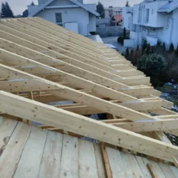 Drewniana konstrukcja dachu w trakcie budowy, widoczne elementy więźby i deskowania, w tle zabudowa jednorodzinna w zimowej scenerii.