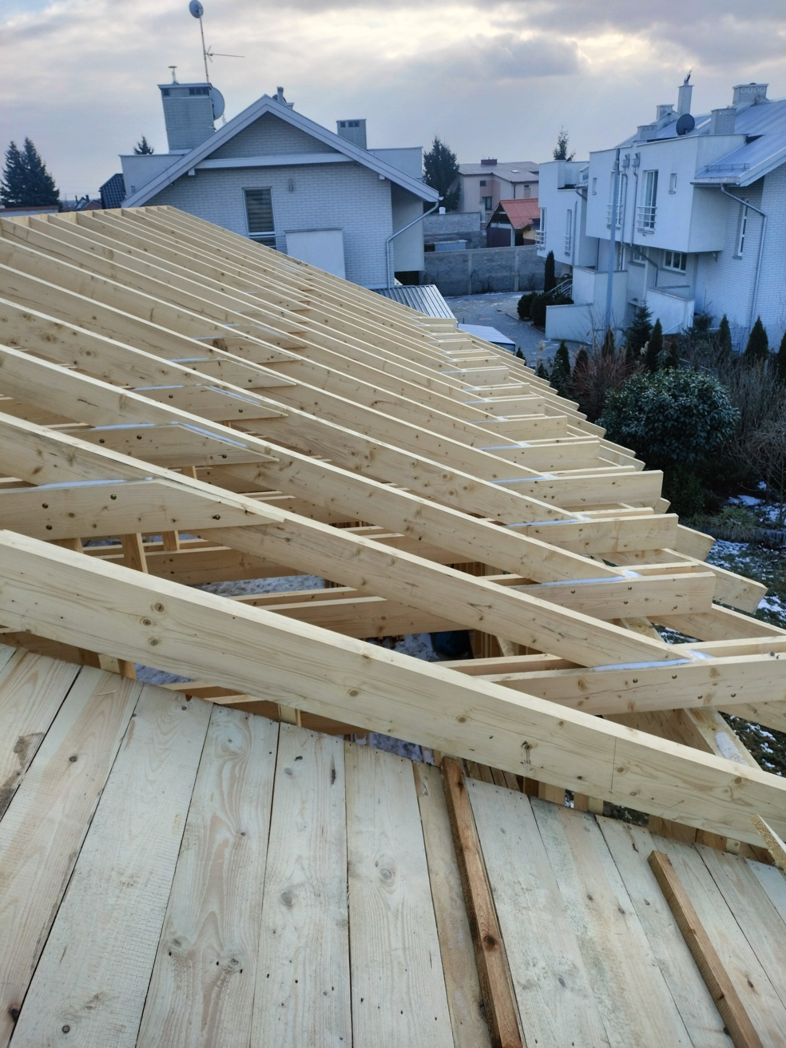 Drewniana konstrukcja dachu w trakcie budowy, widoczne elementy więźby i deskowania, w tle zabudowa jednorodzinna w zimowej scenerii.