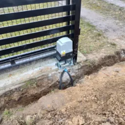 Szara automatyka do bramy przesuwnej zamontowana na fundamencie, widoczne okablowanie w wykopie ziemnym, brama z czarnych metalowych profili.