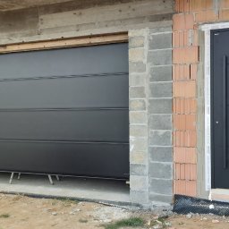 Wojciech Kocoń - Ciemnoszara brama segmentowa garażowa i drzwi wejściowe zamontowane w surowym stanie budynku, widoczne cegły i betonowe nadproże.