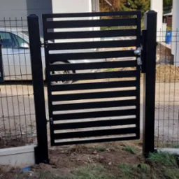 Nowoczesna, czarna furtka z poziomych profili zamontowana w ogrodzeniu z siatki na tle domu jednorodzinnego.