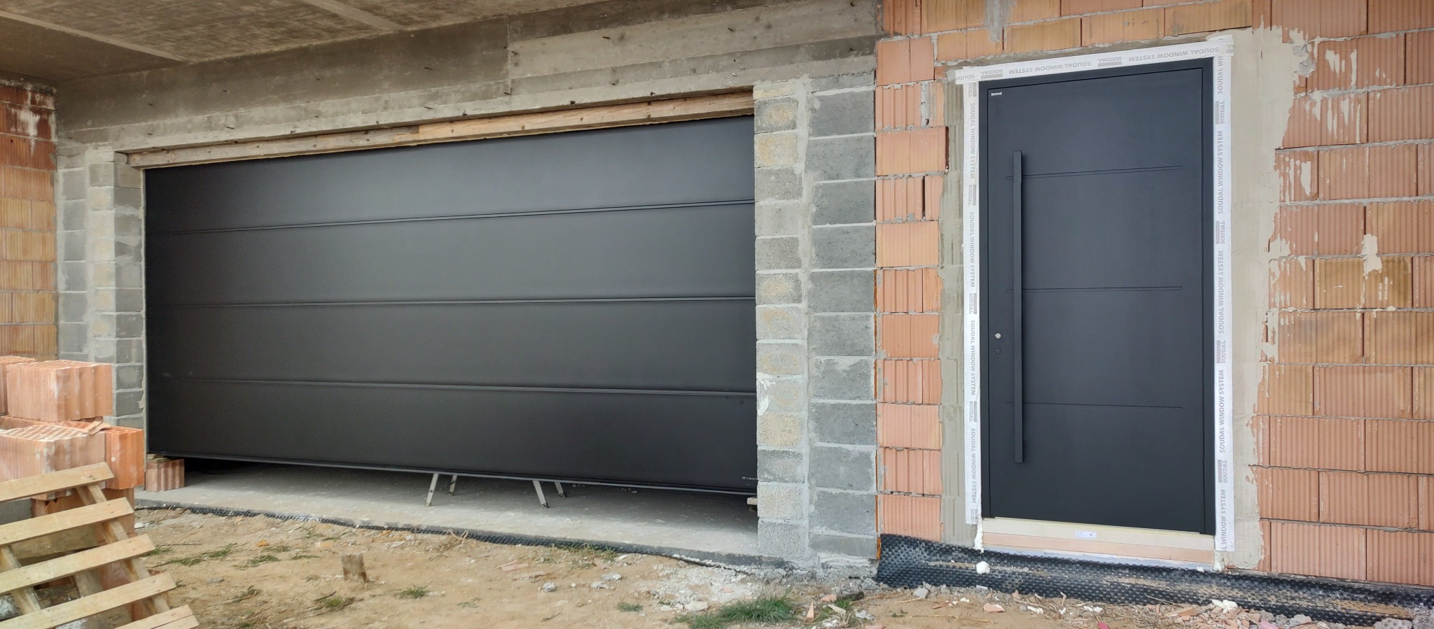 Ciemnoszara brama segmentowa garażowa i drzwi wejściowe zamontowane w surowym stanie budynku, widoczne cegły i betonowe nadproże.
