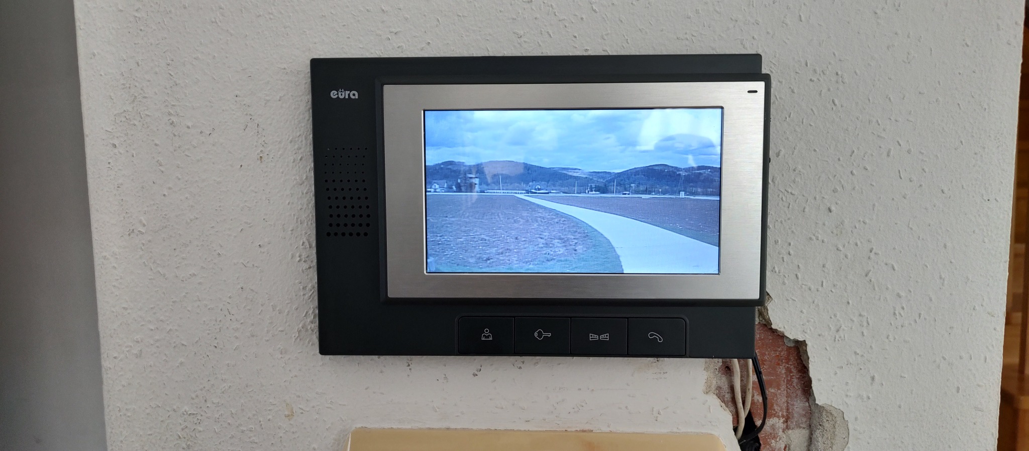 Zamontowany wideofon marki eura z widokiem na zewnątrz budynku, fragment ściany z uszkodzonym tynkiem w tle.