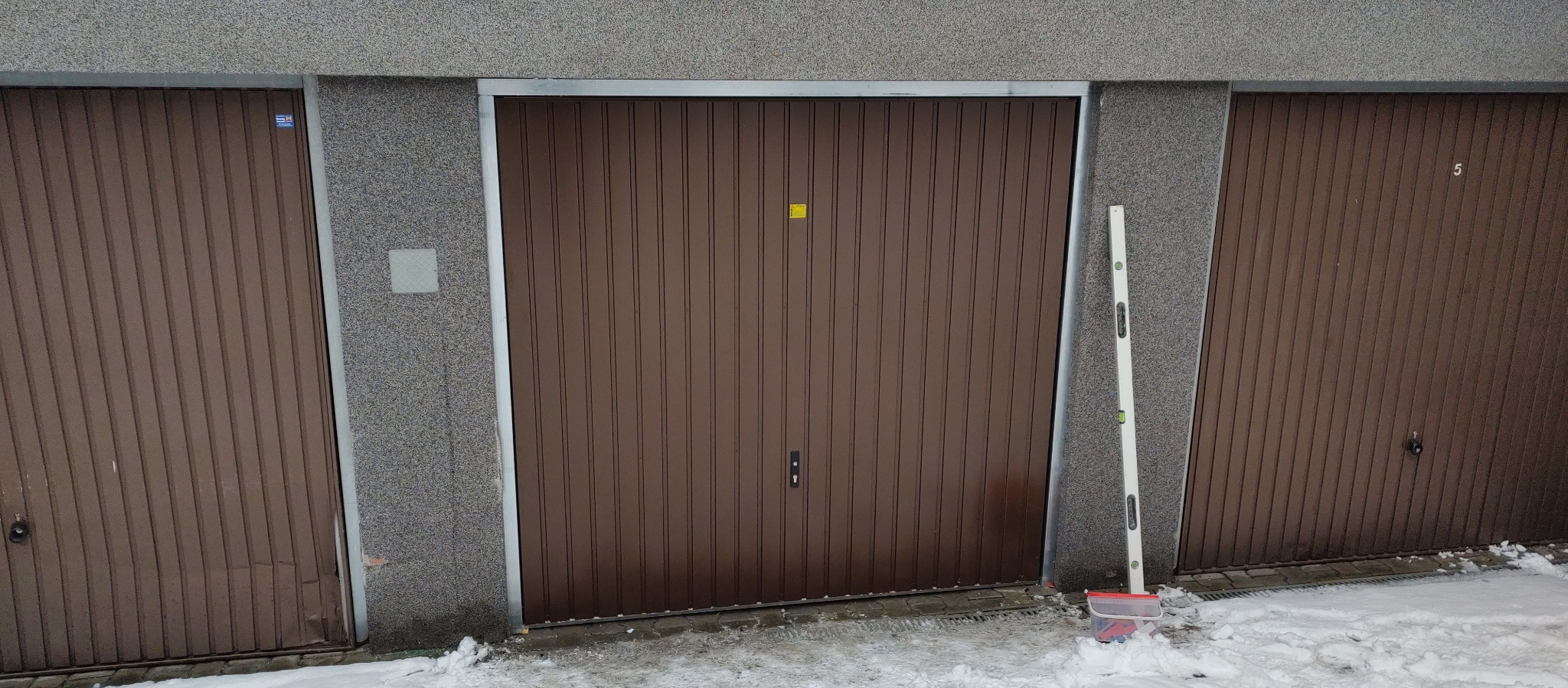 Brązowa brama garażowa segmentowa z pionowymi przetłoczeniami, obok poziomica oparta o ścianę, na ziemi śnieg.