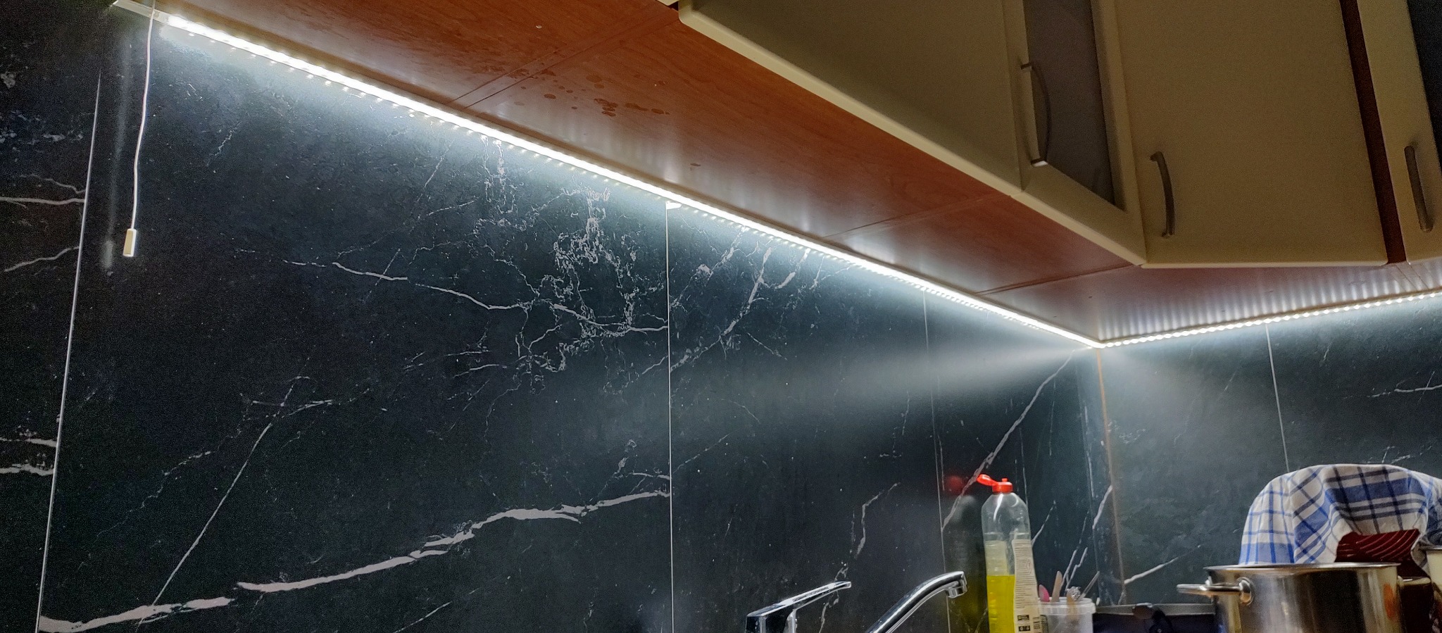 Kuchnia z czarnymi marmurkowymi płytkami i oświetleniem LED podszafkowym, widoczny fragment szafki wiszącej i zlewozmywak.