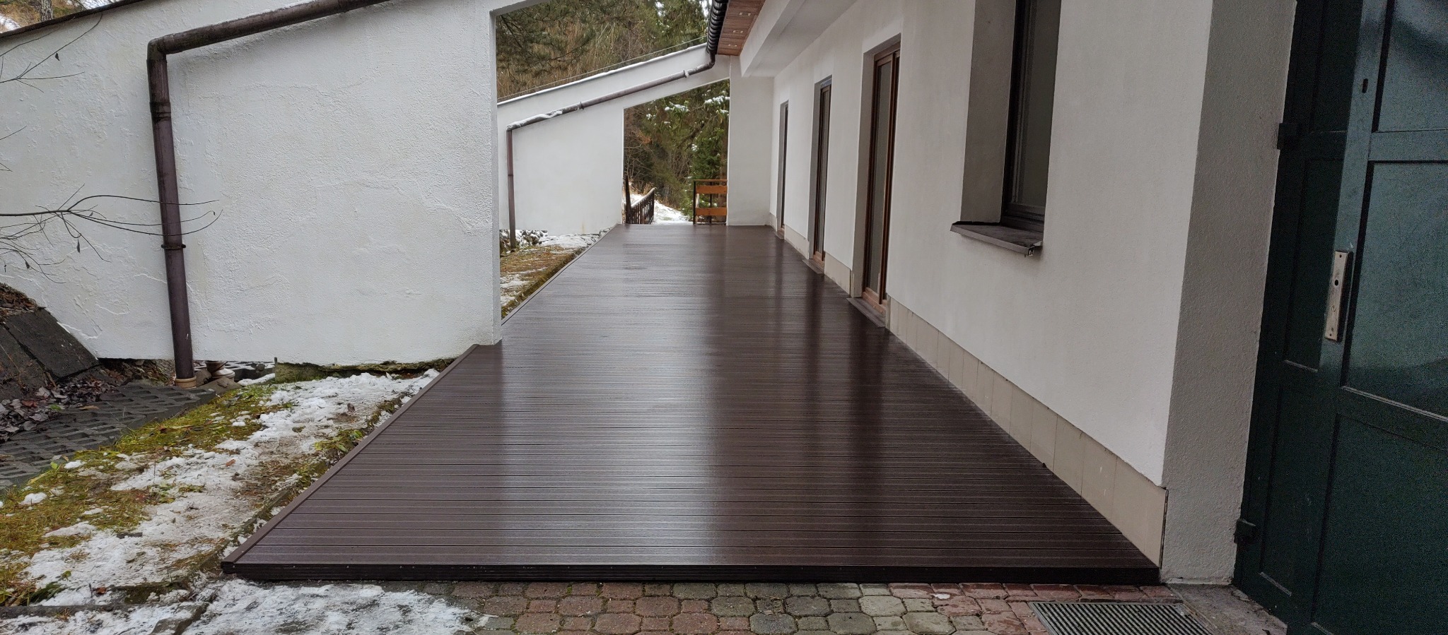 Długi, brązowy taras kompozytowy przylegający do białego budynku z kilkoma wejściami, widoczne resztki śniegu na trawie i chodniku.