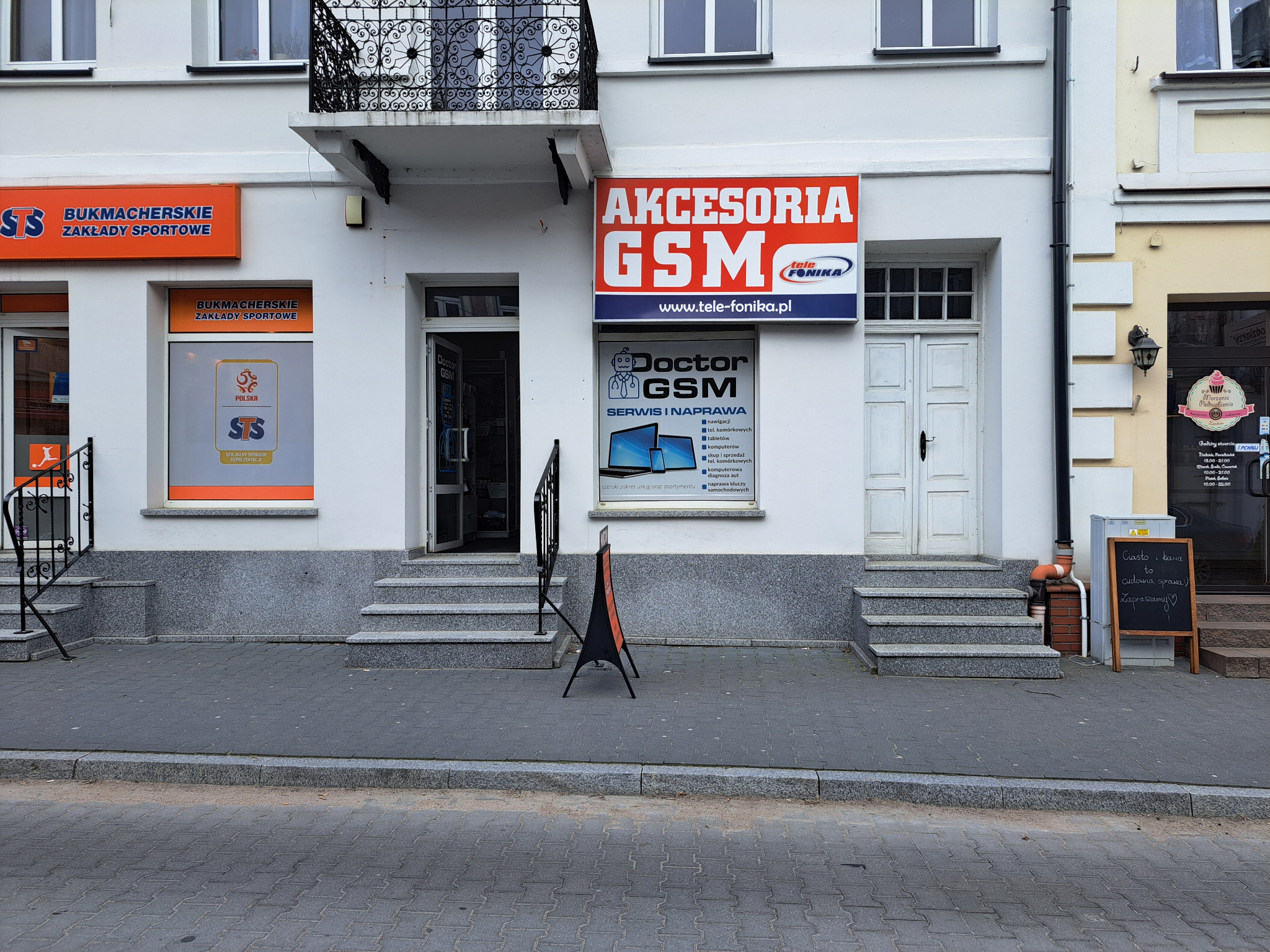 Fasada budynku z szyldami 'Akcesoria GSM', 'Doctor GSM Serwis i Naprawa' oraz 'Bukmacherskie Zakłady Sportowe', widok z ulicy.