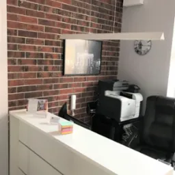 Białe biurko recepcyjne z kalendarzem, karteczkami samoprzylepnymi i drukarką w tle, ściana z cegły i obraz z Bramą Brandenburską.