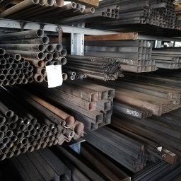 ogrodzenia, wyroby hutnicze, sklep spawalniczy - Ecolux - Magazyn stali: Rzędy rur okrągłych i profili kwadratowych na półkach, widoczne oznaczenia wymiarów na niektórych elementach.