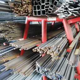 ogrodzenia, wyroby hutnicze, sklep spawalniczy - Ecolux - Różne profile stalowe, pręty okrągłe i kwadratowe, oraz rury prostokątne na czerwonym regale magazynowym.