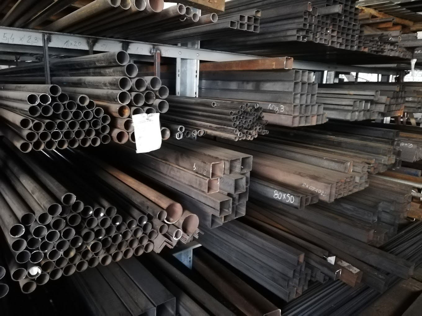 Magazyn stali: Rzędy rur okrągłych i profili kwadratowych na półkach, widoczne oznaczenia wymiarów na niektórych elementach.