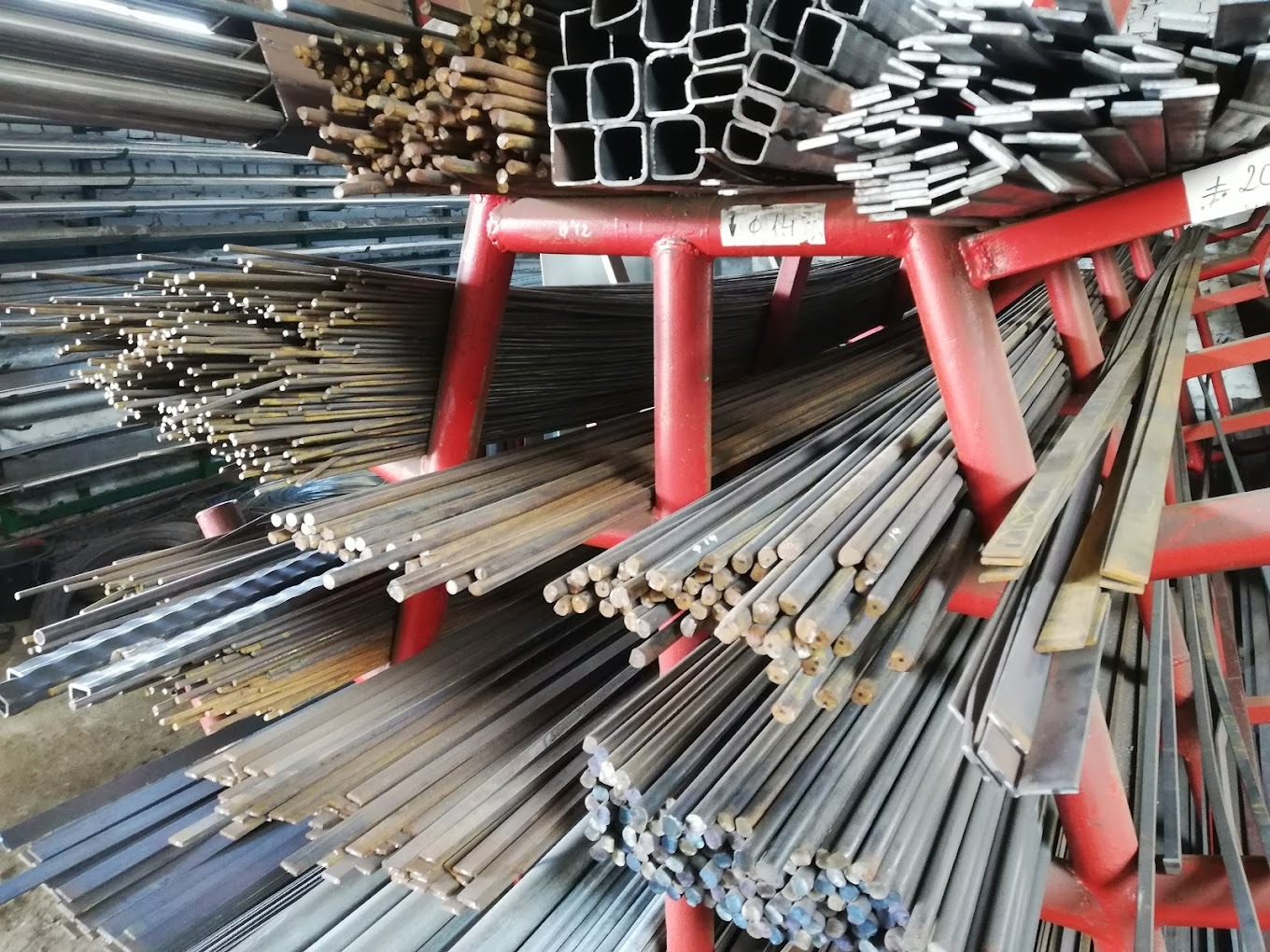 Różne profile stalowe, pręty okrągłe i kwadratowe, oraz rury prostokątne na czerwonym regale magazynowym.