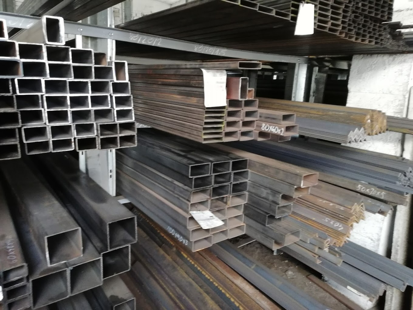 Składowane profile stalowe o różnych przekrojach, w tym kwadratowe i kątowniki, na metalowych regałach w magazynie.
