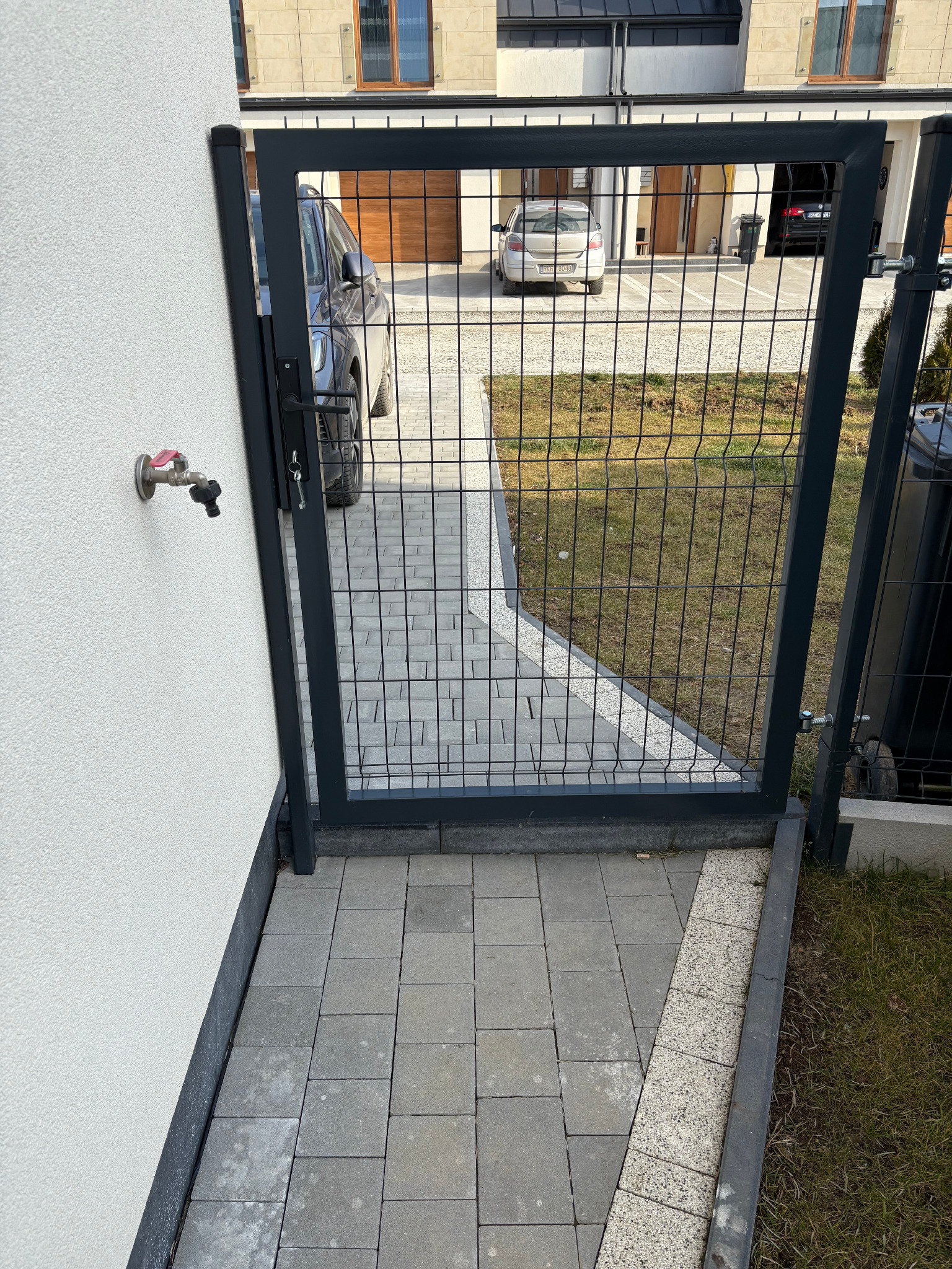 Otwarta furtka z paneli ogrodzeniowych w kolorze antracytowym, prowadząca na posesję wyłożoną szarą kostką brukową, z widocznym zaworem zewnętrznym na ścianie budynku.