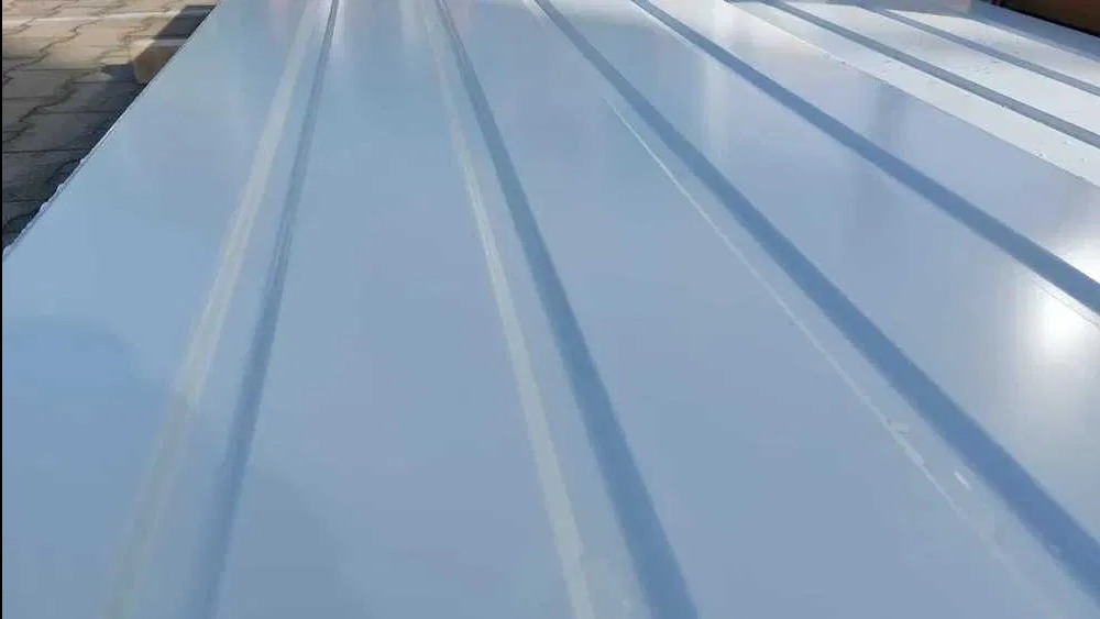 Jasnoszara blacha trapezowa z wyraźnymi przetłoczeniami, ułożona w stos. Widoczne refleksy światła na powierzchni, sugerujące świeże malowanie.