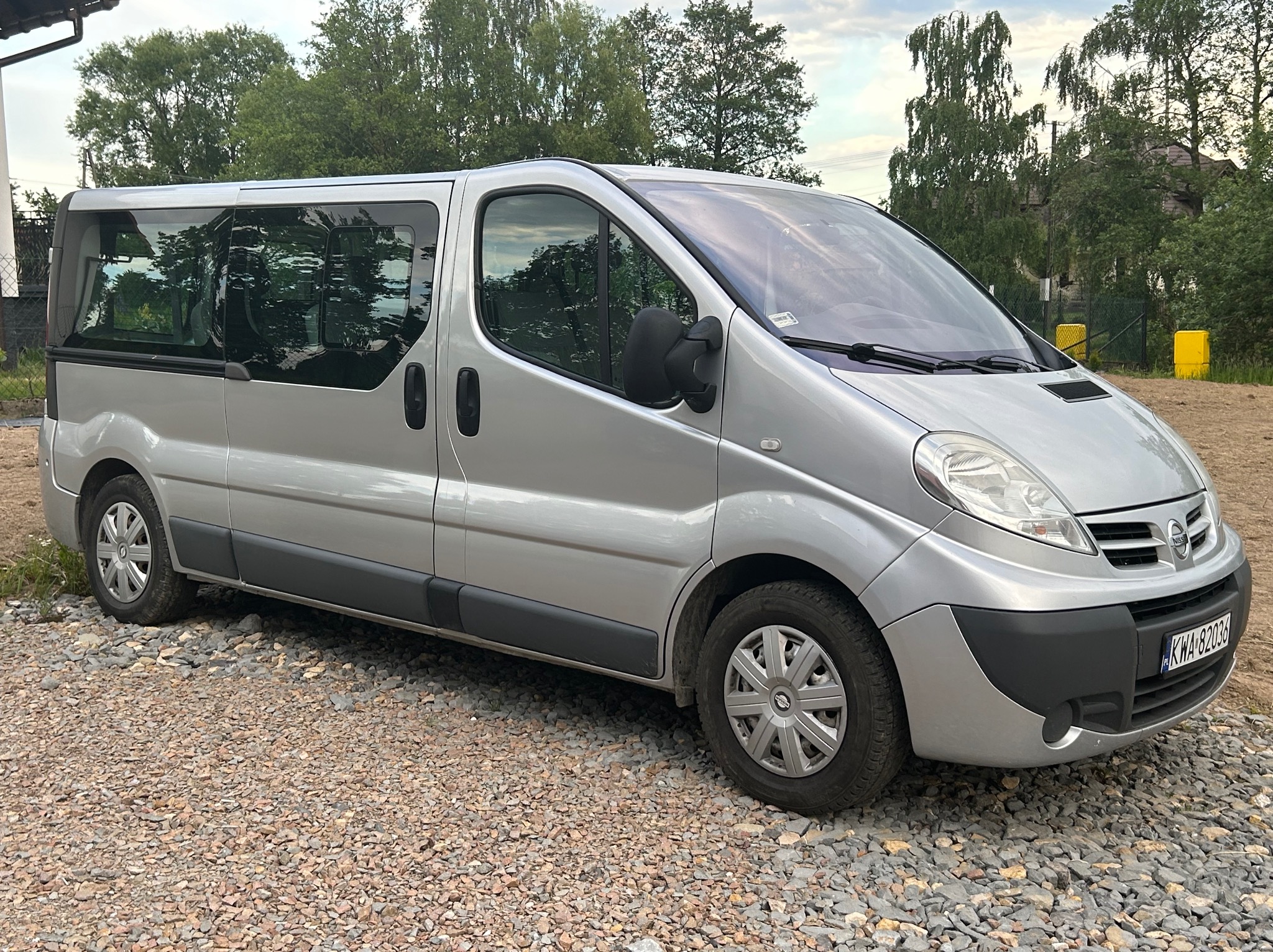 Srebrny van Nissan Primastar zaparkowany na żwirowym podłożu, widok z boku, tablica rejestracyjna widoczna.