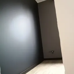 Pomieszczenie po malowaniu ścian na kolor grafitowy, widoczna listwa przypodłogowa w kolorze czarnym oraz jasna podłoga imitująca drewno, gniazdko elektryczne na szarej ścianie.