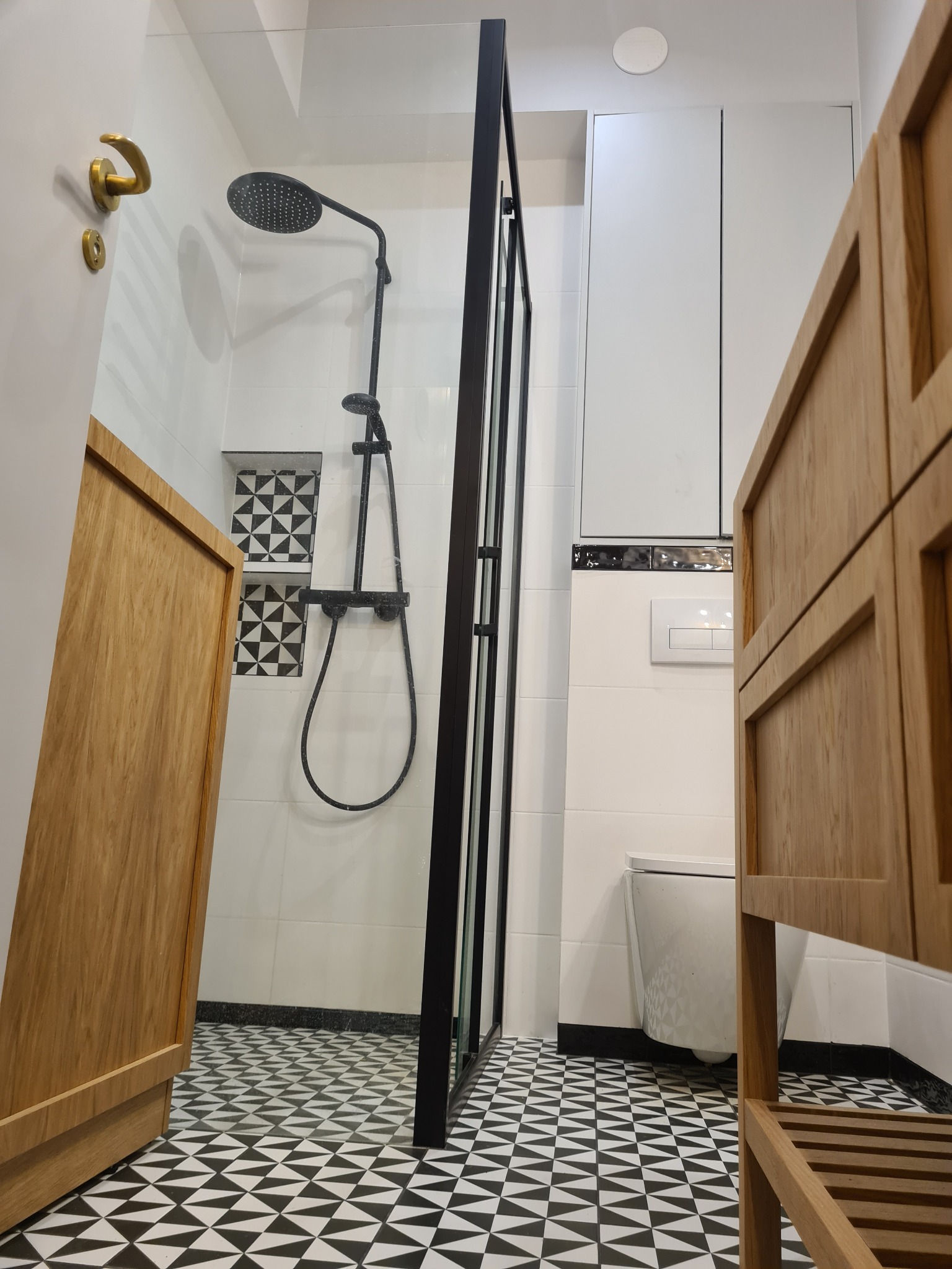 Nowoczesna łazienka z kabiną prysznicową typu walk-in, czarno-białą mozaiką na podłodze i wnęką, oraz drewnianą szafką z półkami po prawej stronie, widok z dołu.