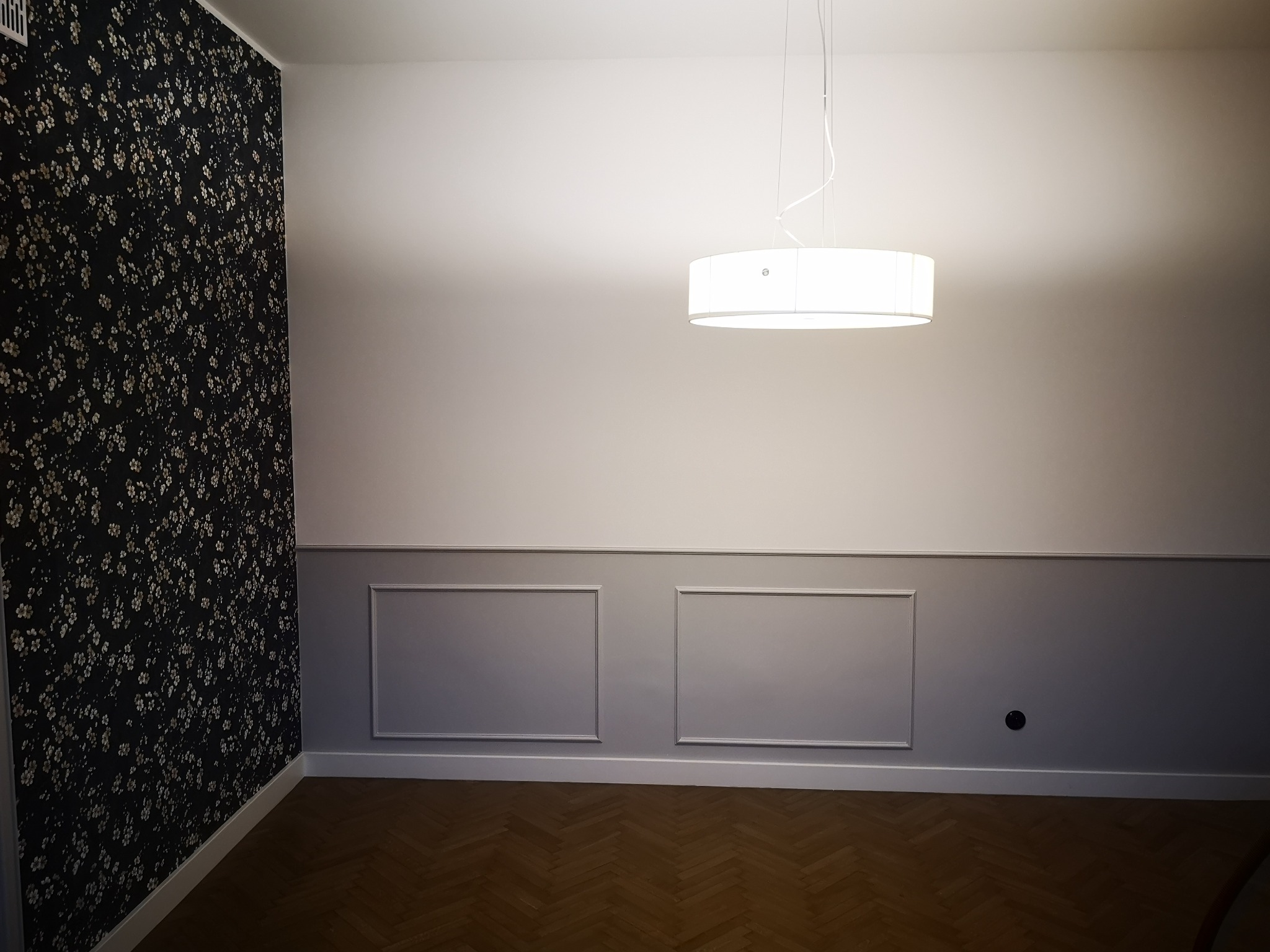 Pusty pokój po remoncie z parkietem w jodełkę, ścianą z tapetą w kwiaty, szarą boazerią i nowoczesną lampą sufitową.