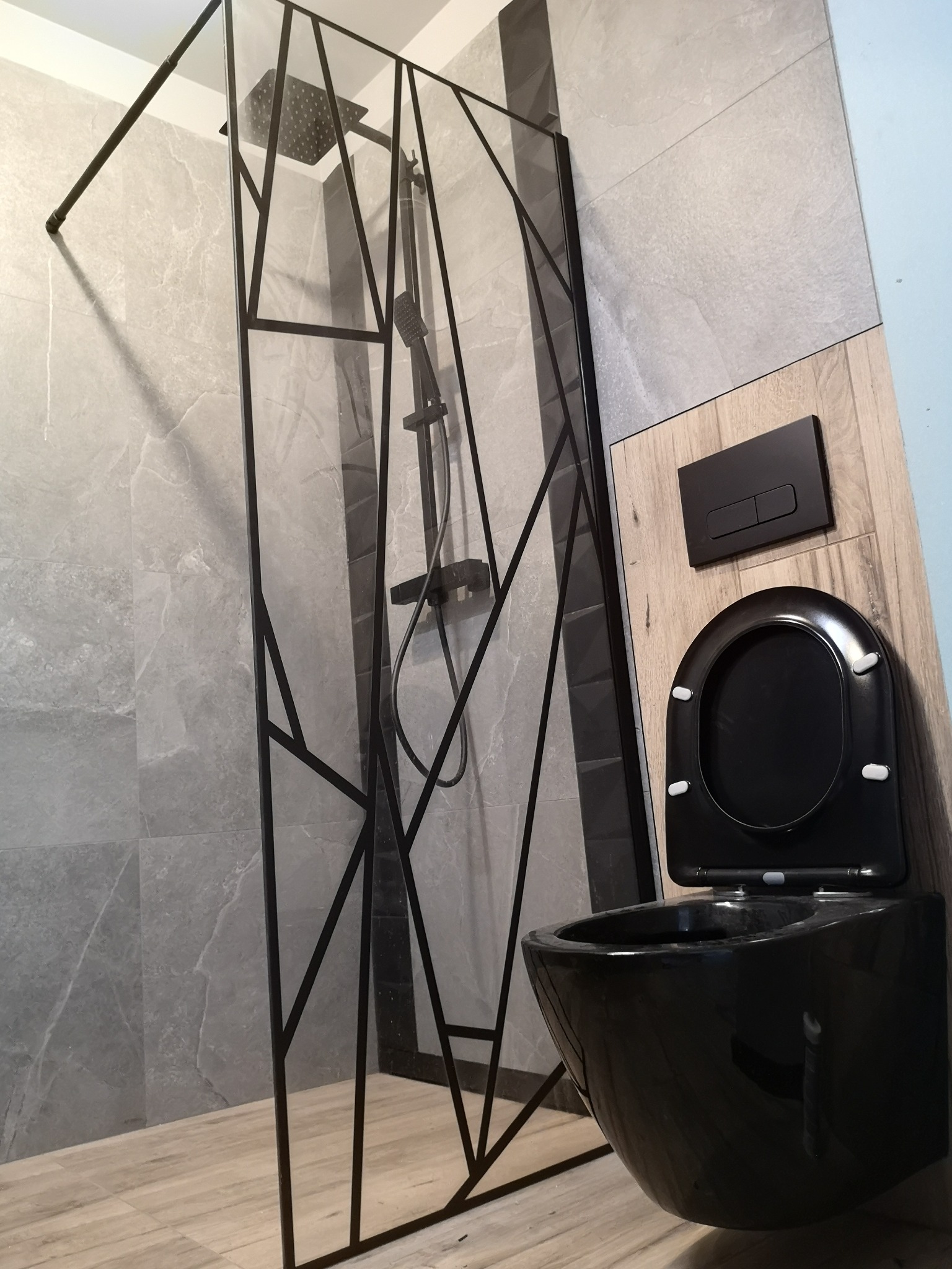 Nowoczesna łazienka z kabiną prysznicową o geometrycznym designie i podwieszaną, czarną toaletą z przyciskiem spłukującym w kolorze czarnym, płytki imitujące marmur na ścianach.