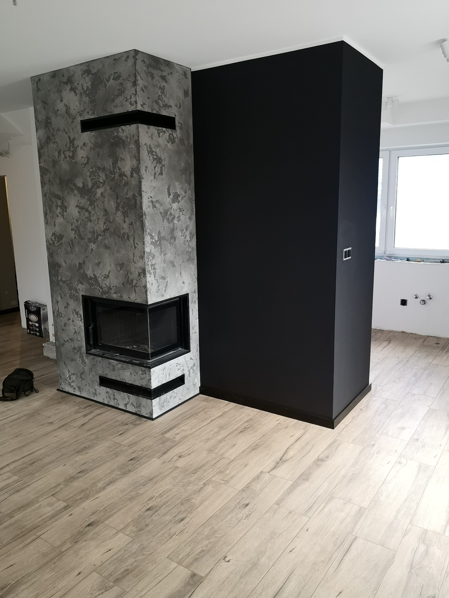 Nowoczesny salon z wykończonym kominkiem o szarej, teksturowanej powierzchni i czarnymi akcentami, kontrastujący z czarną ścianą i podłogą w panelach imitujących jasne drewno.