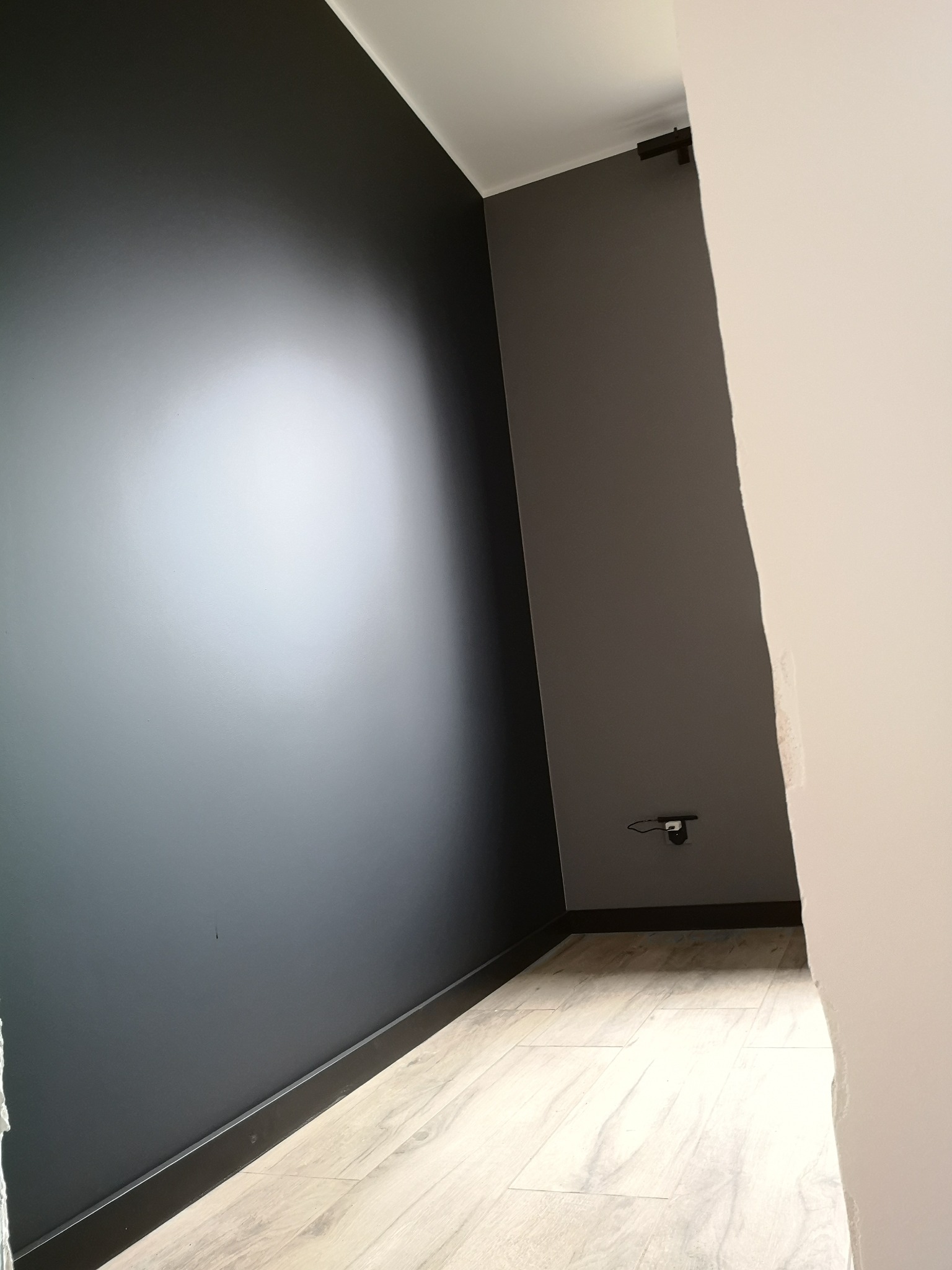 Pomieszczenie po malowaniu ścian na kolor grafitowy, widoczna listwa przypodłogowa w kolorze czarnym oraz jasna podłoga imitująca drewno, gniazdko elektryczne na szarej ścianie.