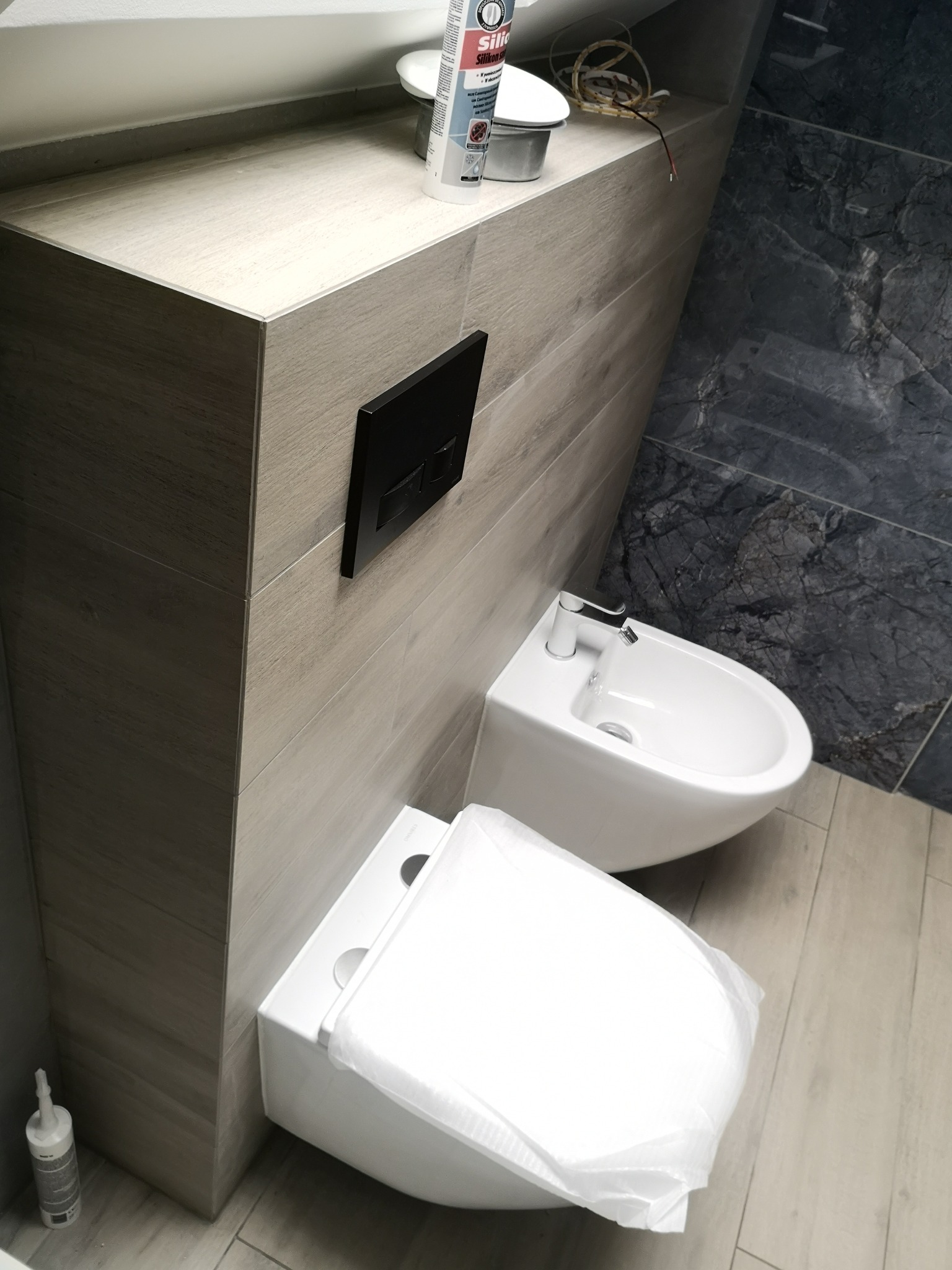 Nowoczesna łazienka w Kobyłce po remoncie: biała toaleta i bidet na tle szaro-beżowych płytek imitujących drewno, z akcentem ciemnego marmuru na ścianie i czarnym przyciskiem spłuczki.