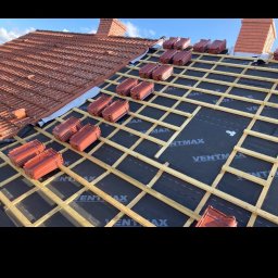 Artur Novikau - Układanie dachówki na łatach pokrytych membraną. Widok na dach z częściowo ułożonym pokryciem, kominem i błękitnym niebem. Prace dekarskie w toku.