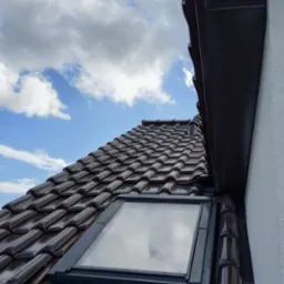 Błyszczący, brązowy dach z dachówką ceramiczną i oknem dachowym, widziany pod kątem. W tle błękitne niebo z chmurami i fragment rynny.