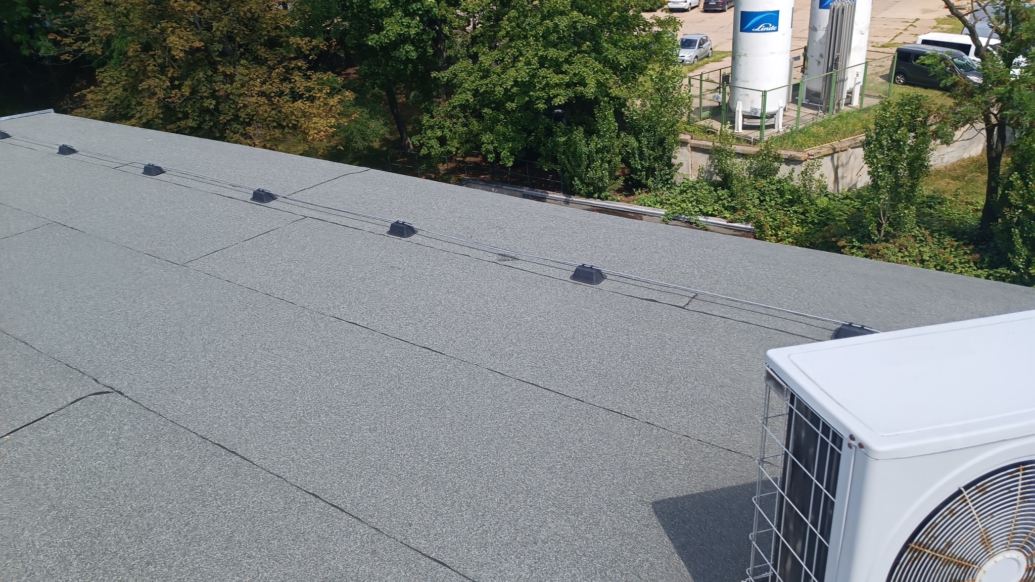 Szary dach pokryty papą termozgrzewalną z elementami mocującymi i klimatyzatorem w rogu, na tle zieleni i budynków przemysłowych.