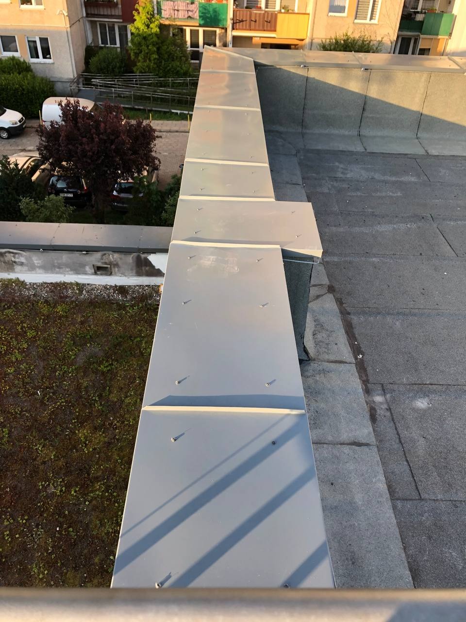 Szara obróbka blacharska na dachu budynku wielorodzinnego, widok z góry. Elementy mocowane śrubami, w tle zieleń i zabudowa miejska.