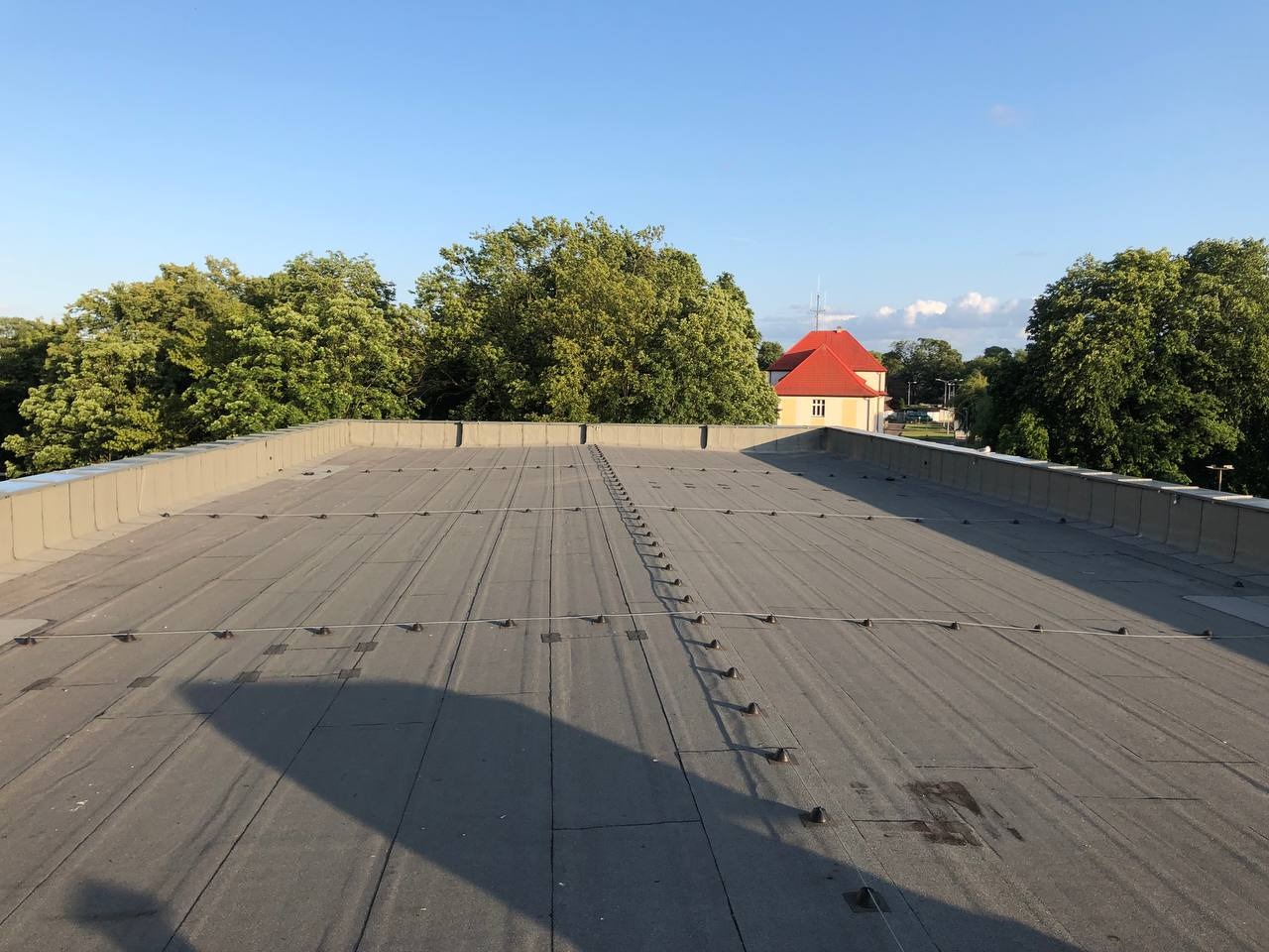 Płaski, szary dach pokryty papą z widocznymi liniami mocowań i elementami blacharskimi na krawędziach. W tle budynek z czerwonym dachem i zieleń drzew.