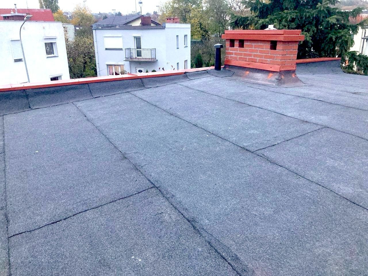 Szary dach pokryty papą z widocznym kominem z czerwonej cegły. W tle budynki mieszkalne i drzewa. Widok z góry na płaską powierzchnię.