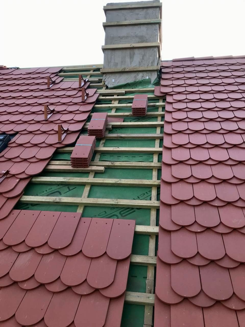 Częściowo pokryty dach czerwoną dachówką karpiówką, widoczna konstrukcja drewniana i komin. Ułożone stosy dachówek czekają na montaż.