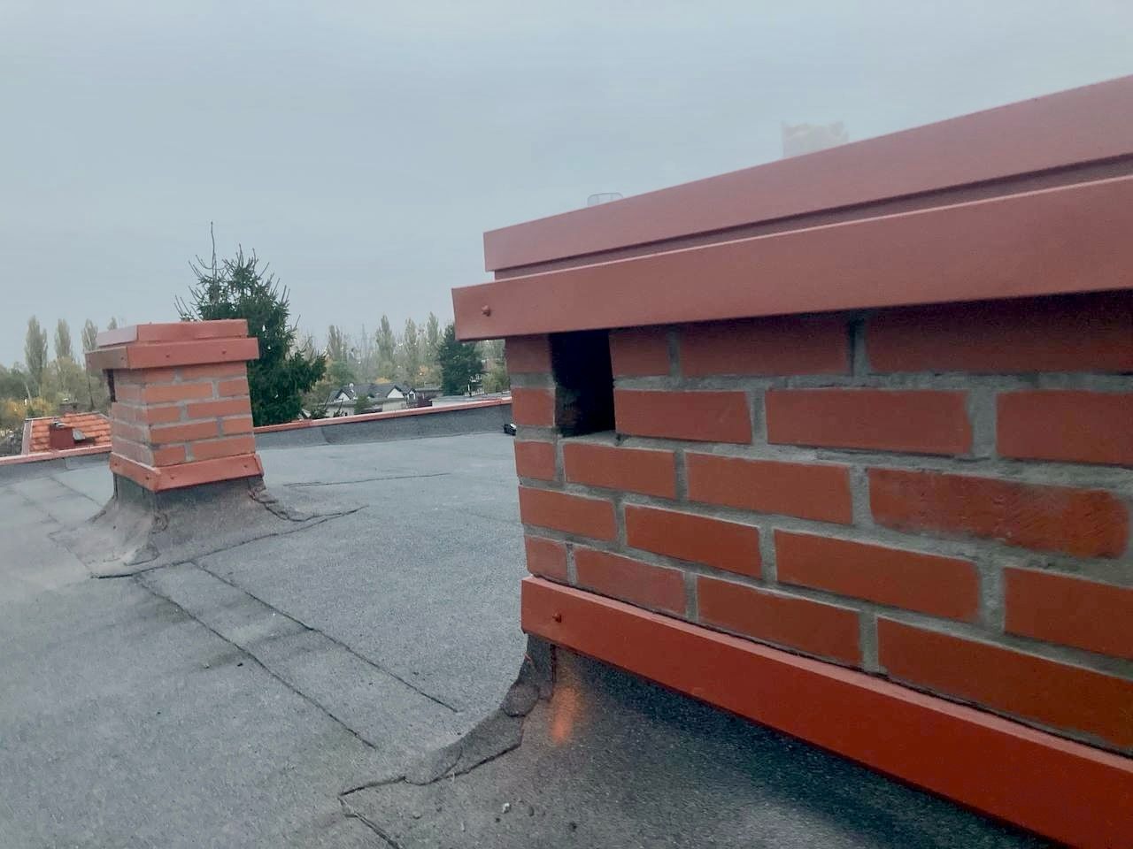 Wykończony komin z cegły z metalową obróbką na dachu pokrytym papą. W tle drugi komin i drzewa. Widok z góry, pochmurny dzień.