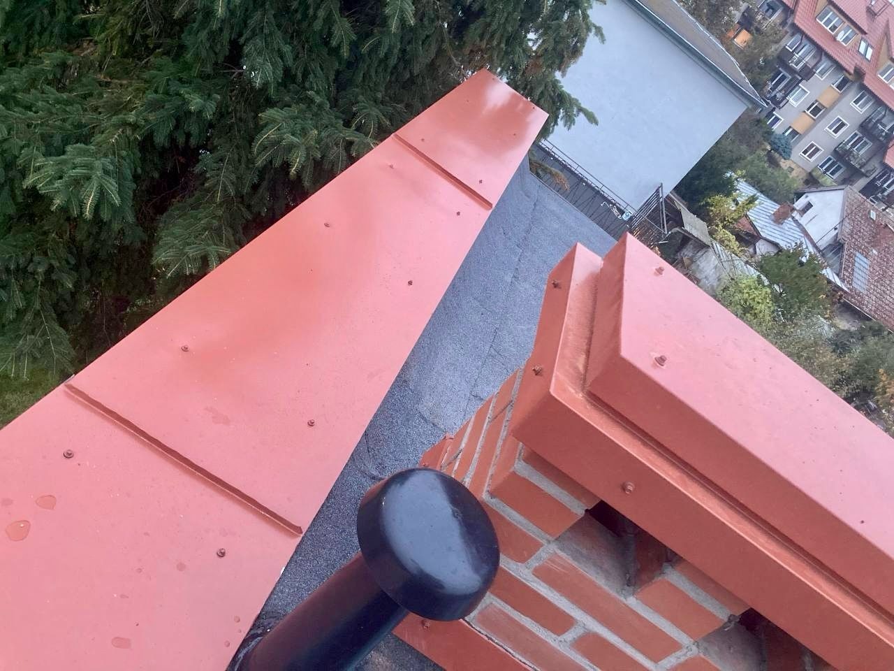 Czerwony dach z blachy na budynku z kominem, widok z góry na tle zieleni i zabudowy miejskiej. Elementy mocowane śrubami.