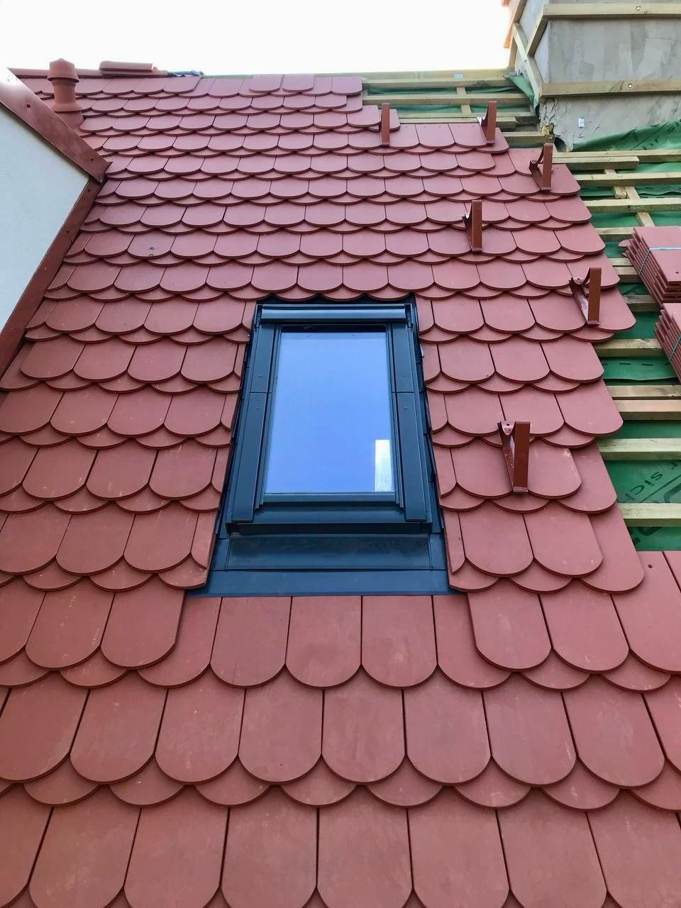 Częściowo pokryty dach czerwoną dachówką karpiówką z oknem dachowym i metalowymi uchwytami, widoczne deski i zielona membrana na niepokrytej części dachu.