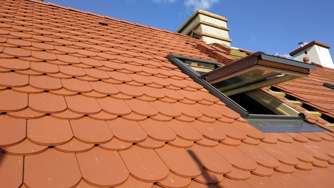 Czerwony dach z dachówki karpiówki, otwarte okno dachowe, widoczne kominy, błękitne niebo. Dachówka ułożona w łuskę. Częściowo widoczne deski pod dachówką.