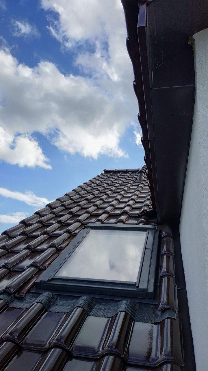 Błyszczący, brązowy dach z dachówką ceramiczną i oknem dachowym, widziany pod kątem. W tle błękitne niebo z chmurami i fragment rynny.