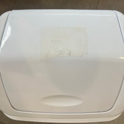 FHU - firma sprzątająca Cleanliness Katarzyna Jeziorna - Biały pojemnik na odpady z resztkami kleju po naklejce na pokrywie, widok z góry na brązowe płytki podłogowe.