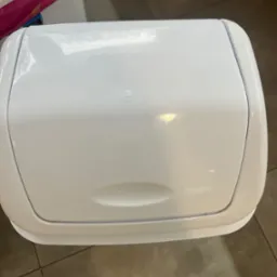 Zamknięty, biały pojemnik na pieluchy na brązowych płytkach podłogowych, widziany z góry.