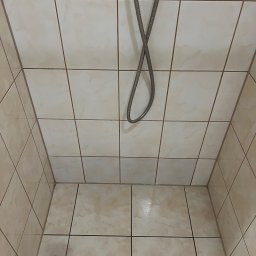 FHU - firma sprzątająca Cleanliness Katarzyna Jeziorna - Kabiną prysznicowa wyłożona beżowymi, kwadratowymi płytkami z delikatnym marmurkowym wzorem, widoczny wąż prysznicowy zwisający z góry.