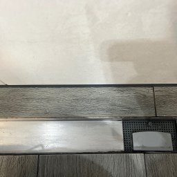 FHU - firma sprzątająca Cleanliness Katarzyna Jeziorna - Szczegół odpływu liniowego prysznicowego z szarą kratką i napisem 'PULL OUT', wkomponowanego w płytki imitujące drewno, z widoczną górną częścią ściany wyłożonej jasnymi płytkami.